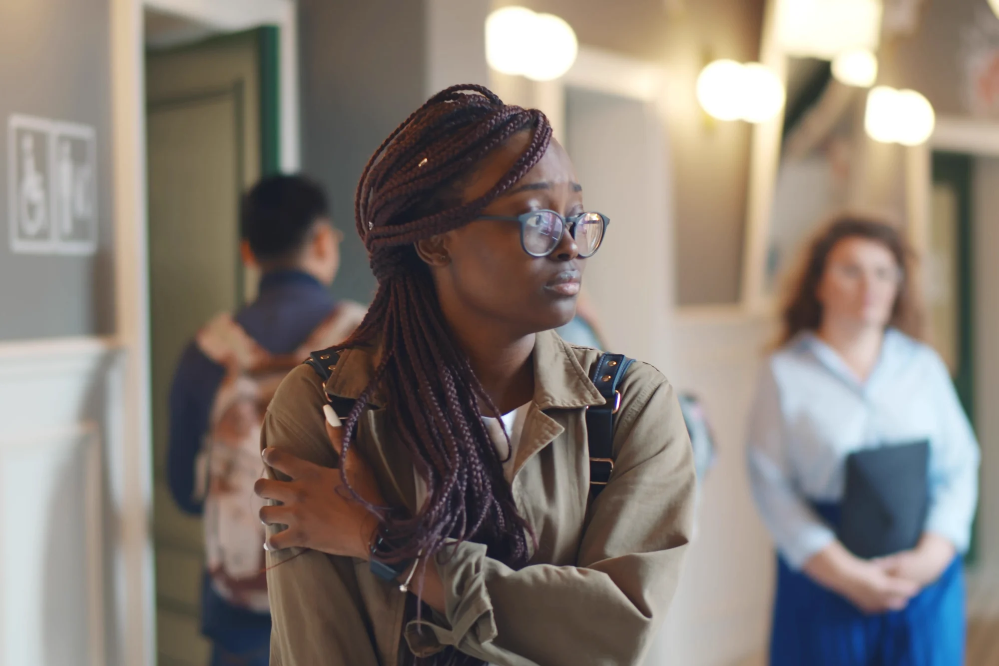 A woman with braided hair and glasses, wearing a khaki jacket, stands in a college corridor with a worried expression, clutching her arms tightly.