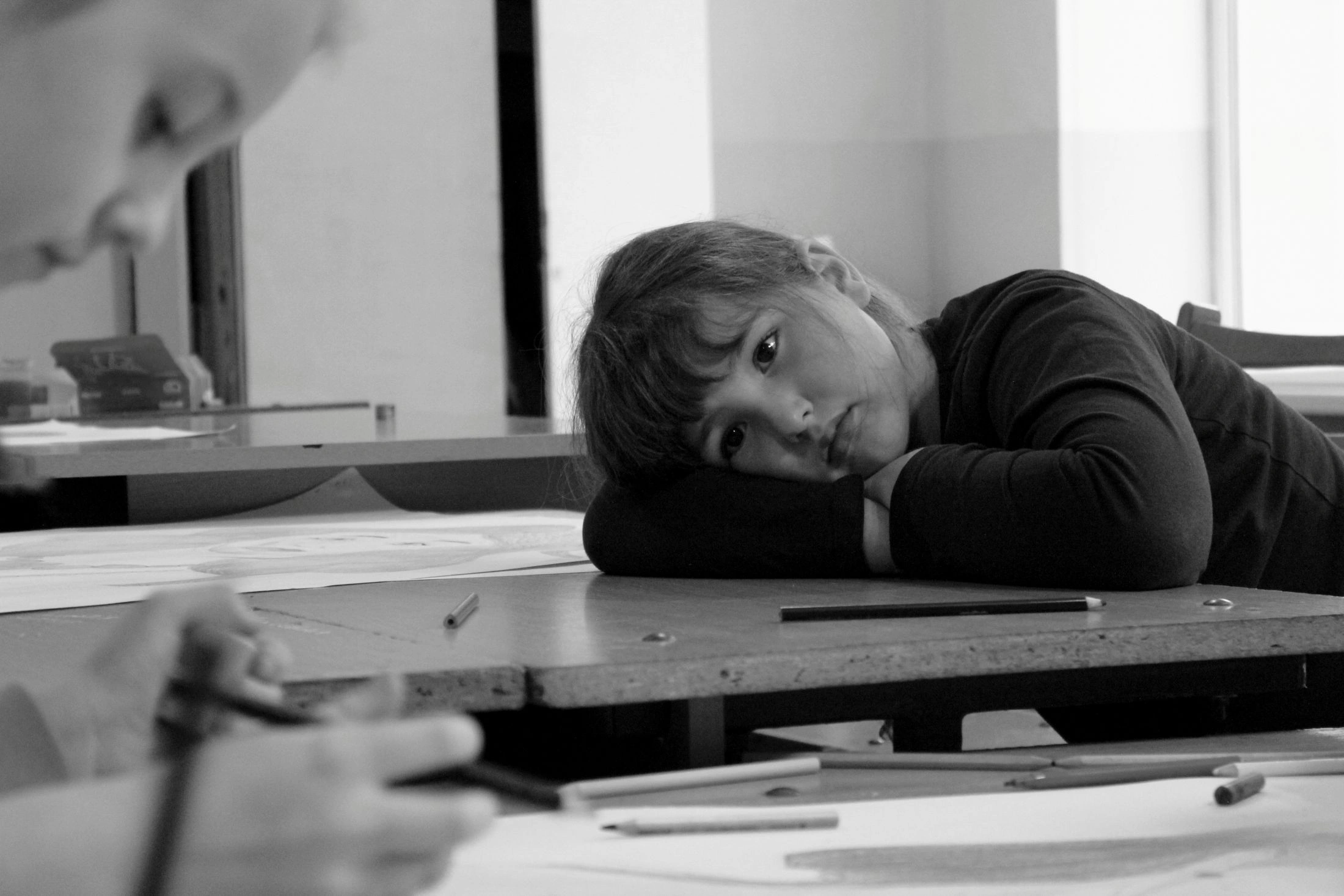 A black and white photo of a young girl resting her head on her arms on a wooden desk, looking directly at the viewer with a somber expression.