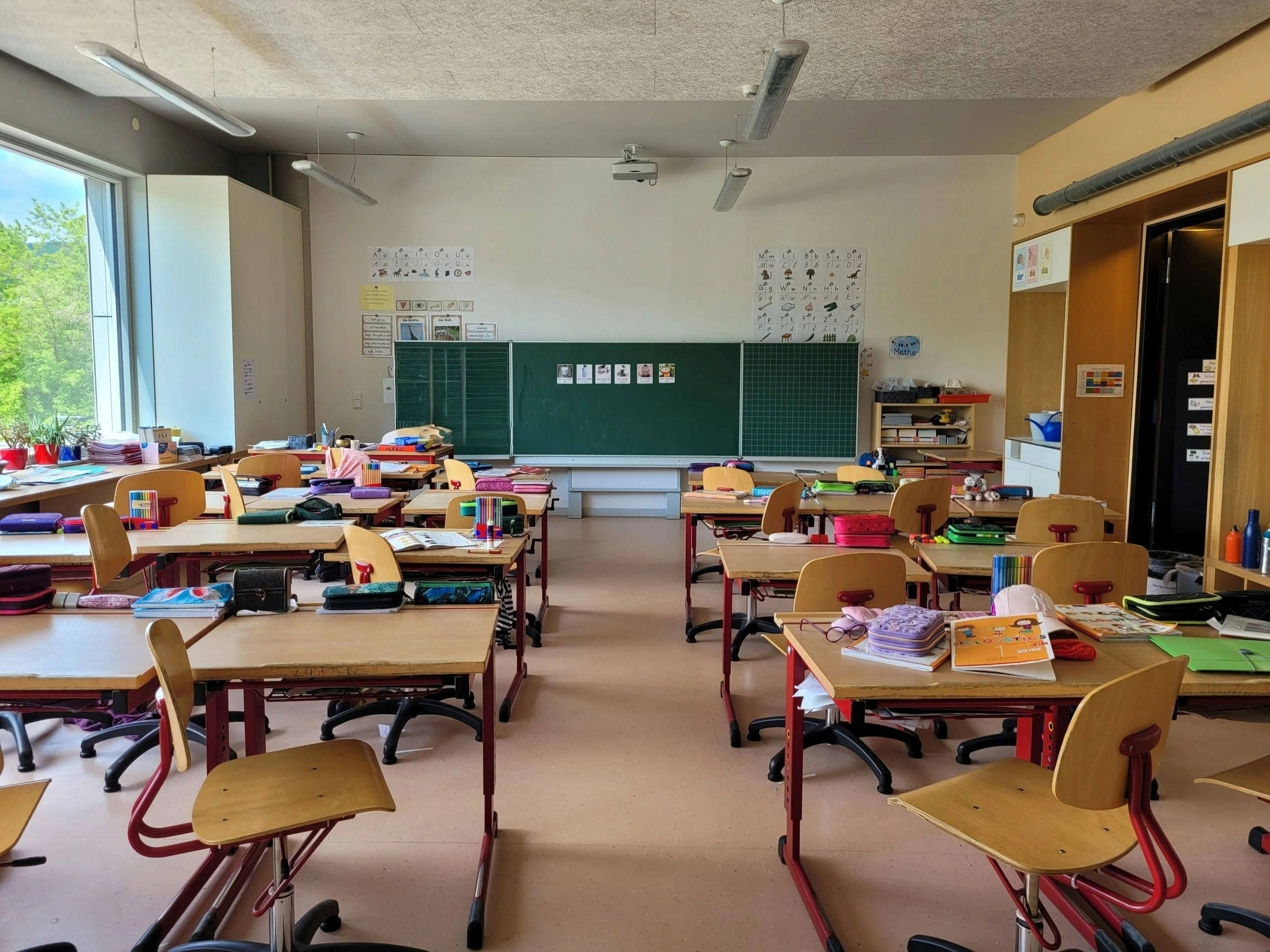An empty, modern classroom with rows of small wooden desks and chairs, facing a large green blackboard covered in academic posters. Supplies are ready on the desks.