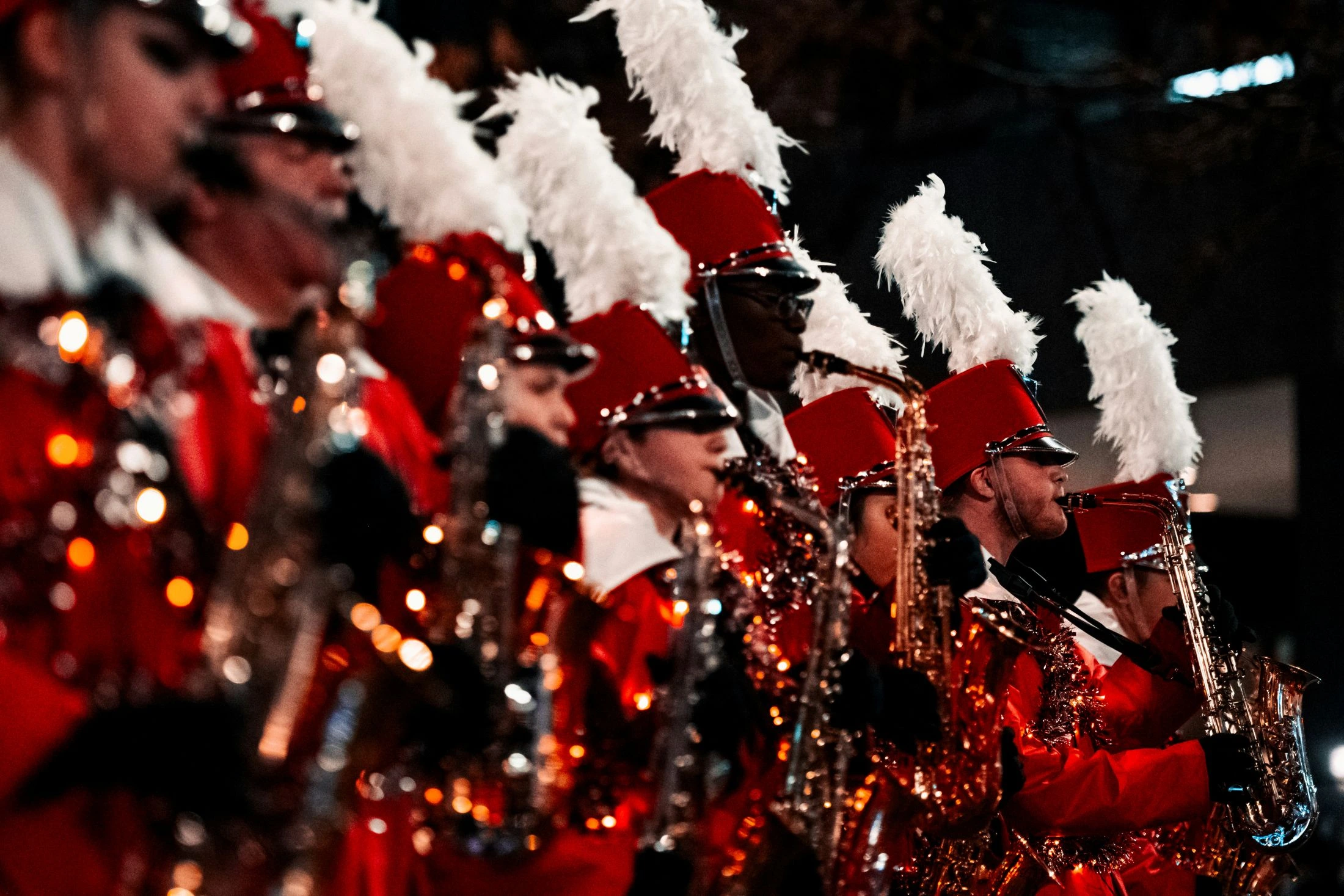 Members of a marching band in red uniforms and plumed hats play saxophones during a night performance. Instruments reflect bright lights.