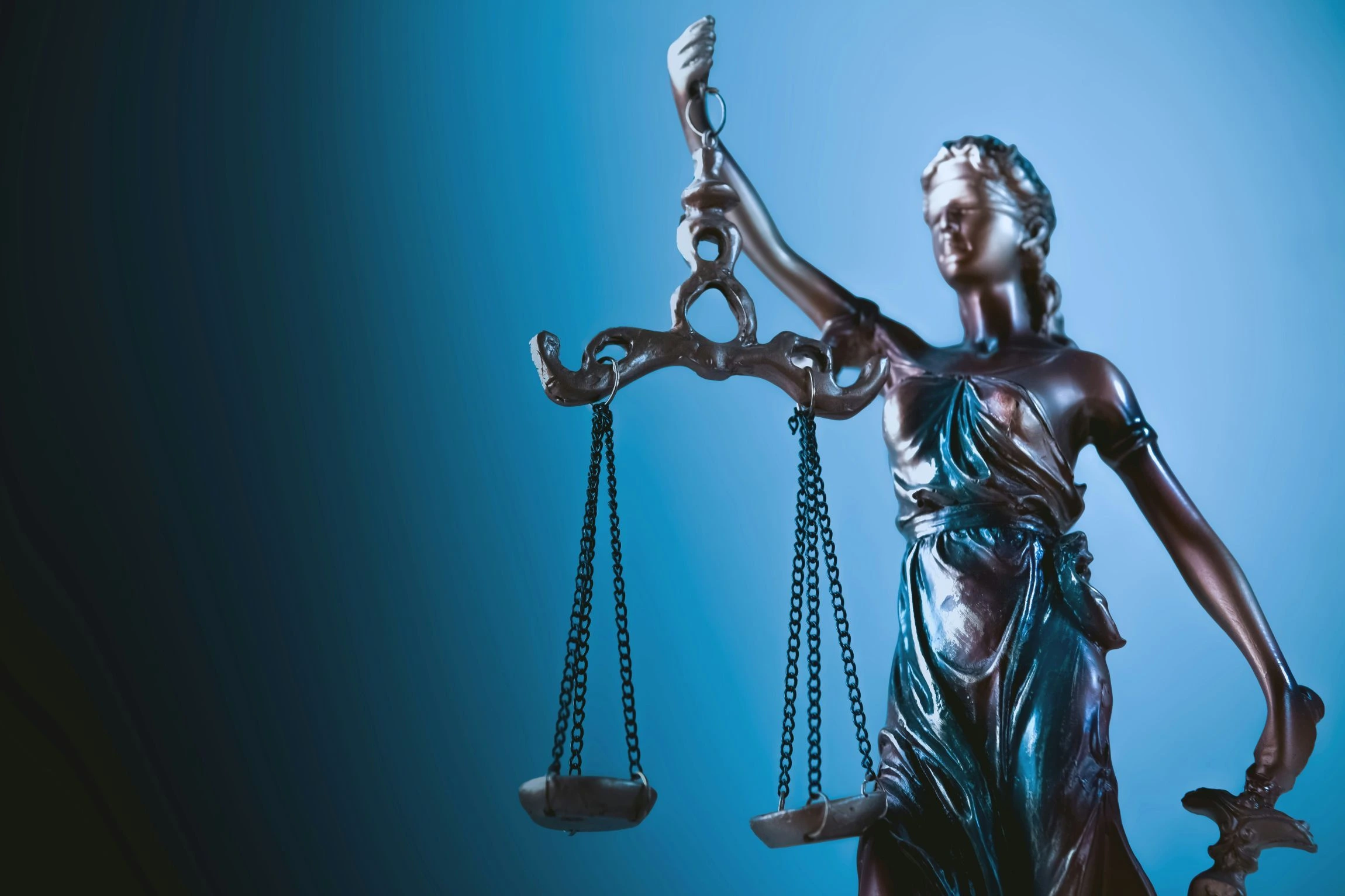A bronze statue of Lady Justice, blindfolded, holds the balanced scales of justice in her raised hand against a dramatic blue background.