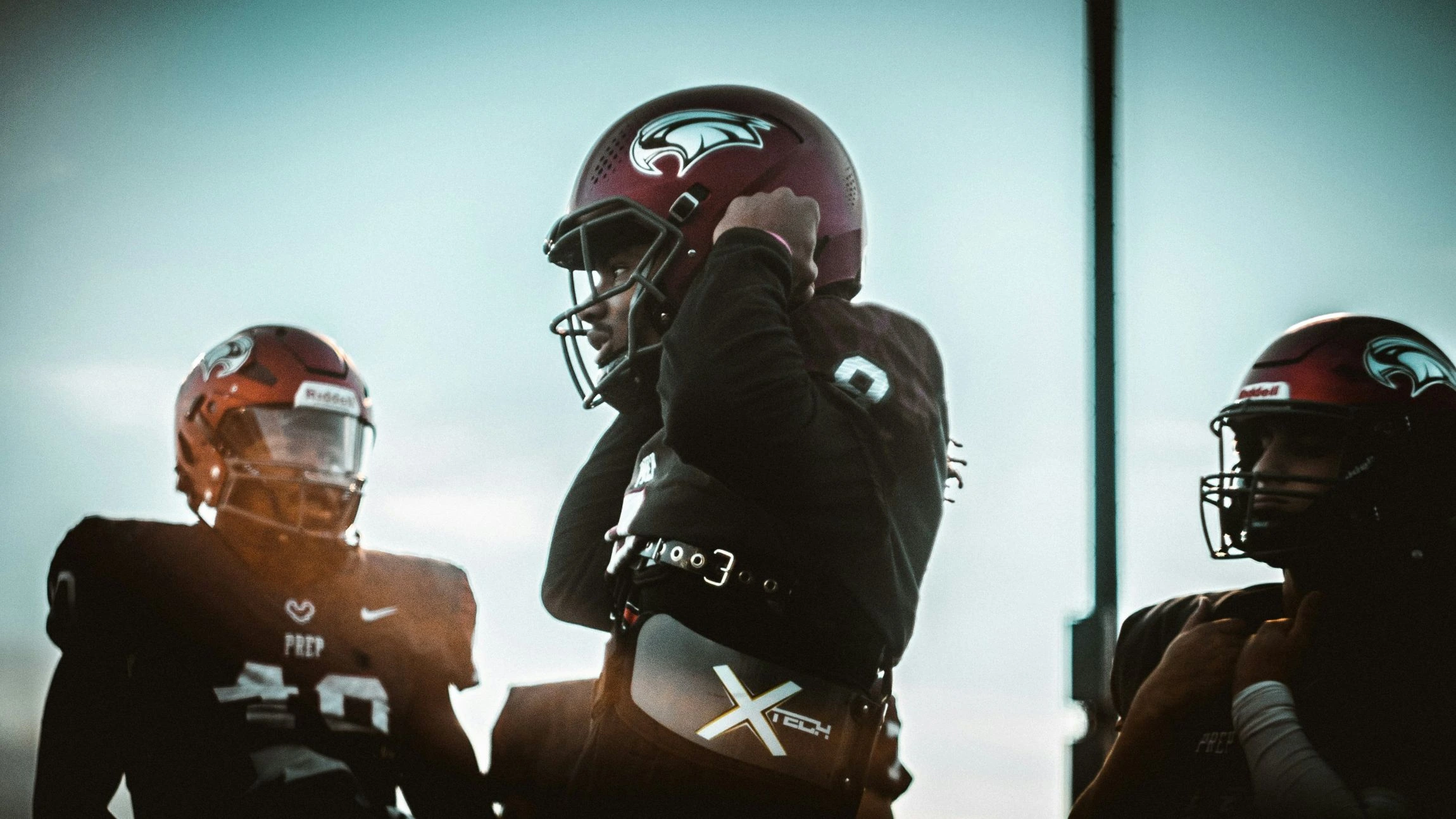 Three football players stand silhouetted against a bright sky. The player in the foreground adjusts his maroon helmet and wears a visible protective belt.